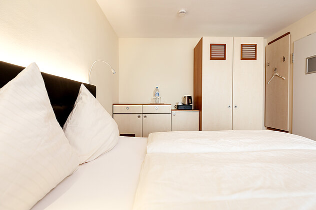 Modernes Hotelzimmer mit gemachter weißer Bettwäsche, Kommode, Wasserkocher, Kleiderschrank und Wandbeleuchtung.