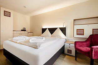Modernes Doppelzimmer mit weiß aufbereitetem Bett, zusammengelegten Handtüchern und einem roten Sessel.