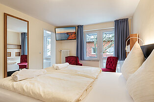 Blick vom Doppelbett auf einen Wandspiegel, Flachbild-Fernseher und ein Fenster mit blauen Vorhängen.