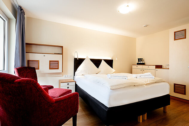 Gemütliches Hotelzimmer mit gemachtem Doppelbett, zwei samtigen roten Sesseln und heller Holzoberfläche.